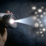 Eine Hand hält eine Kamera, aus deren Objektiv ein Lichtstrahl auf einen dunklen Hintergrund mit zahlreichen leuchtenden Symbolen für digitale Medien und Wiedergabe projiziert wird.