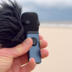 Eine Hand hält ein kleines blaues Mikrofon mit einem schwarzen Windschutz an einem Strand.