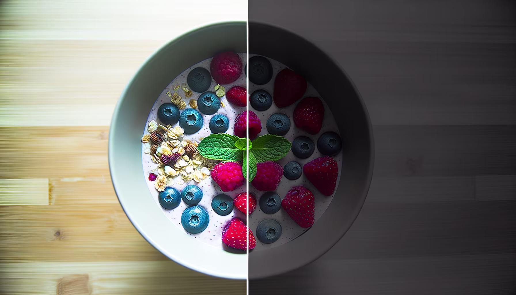 Das Bild zeigt eine Schale mit einem Frühstücks- oder Smoothie-Bowl, die vertikal in zwei Hälften geteilt ist. Die linke Seite präsentiert die Schale in leuchtenden, natürlichen Farben und hellem Licht. Man sieht eine cremige Basis, bestreut mit frischen Blaubeeren, Himbeeren, Erdbeeren und knusprigem Müsli, garniert mit einem Minzblatt. Die Farben der Beeren sind satt und einladend. Die rechte Seite zeigt genau dieselbe Schale, jedoch in einer stark abgedunkelten und entsättigten Version. Die Farben sind matt und gräulich, die Details weniger erkennbar, und der Gesamteindruck ist deutlich weniger appetitlich. Dieses Bild demonstriert eindrucksvoll den Unterschied, den Beleuchtung und Farbsättigung auf die visuelle Wahrnehmung von Speisen haben können. **Alternativtext:** Eine vertikal geteilte Ansicht einer Schale mit Joghurt, Beeren, Müsli und Minze, wobei die linke Hälfte hell und farbenfroh ist und die rechte Hälfte dunkel und entsättigt.