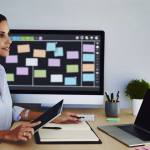 Eine konzentrierte Frau sitzt an einem Schreibtisch, hält ein Tablet und einen Stift über einem Notizbuch, während auf dem Monitor hinter ihr eine digitale Plantafel mit bunten Aufgaben zu sehen ist.