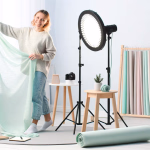 Eine lächelnde Frau hält einen mintgrünen Stoff in einem hellen Kreativstudio, umgeben von professioneller Beleuchtung und einer Auswahl an farbigen Hintergrundstoffen.