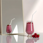 Ein Glas Himbeer-Smoothie mit Strohhalm und frischen Himbeeren, neben einem Spiegel auf einer reflektierenden Oberfläche.