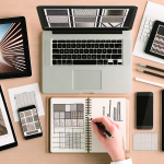 **Vorschlag (Text):** Auf einem hellen Holztisch sind von oben gesehen mehrere digitale Geräte und analoge Designmaterialien ausgebreitet. Ein offener Laptop im Zentrum zeigt eine Reihe von Mustern auf seinem Bildschirm. Links davon liegt ein Tablet mit dem Bild eines modernen Holzgangs, der sich zu einem hellen Punkt verjüngt. Rechts über einer weißen Tastatur befindet sich ein weiteres Tablet, ebenfalls mit einer Auswahl an Texturen. Im unteren Bereich des Tisches ist ein weiteres Tablet mit einer ähnlichen Architekturaufnahme zu sehen, daneben ein Smartphone. Eine Hand zeichnet in einem offenen Notizbuch Muster und Diagramme. Verschiedene Stifte und weitere Papiere sind ebenfalls auf dem Tisch verteilt. --- **Alternativtext:** Ansicht von oben auf einen hellen Holztisch mit einem Laptop, mehreren Tablets und einem Smartphone. Die Bildschirme der Geräte zeigen diverse digitale Muster und Architekturansichten. Eine Hand zeichnet in einem Notizbuch analoge Muster und Diagramme, umgeben von Stiften und Papieren.