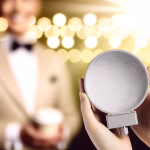 Hände halten einen Mikrofon-Popfilter vor einem unscharfen Sprecher in eleganter Kleidung und festlicher Beleuchtung.