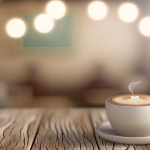 **Vorgeschlagener Text:** Ein Moment der Ruhe und des Genusses: Eine Tasse kunstvoll verzierter Kaffee auf einem Holztisch, im warmen Ambiente eines Cafés. **Alternativtext:** Eine dampfende Tasse Kaffee mit Latte Art auf einem Holztisch in einem gemütlichen Café.