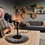 **Vorgeschlagener Text für den Blogbeitrag:** Eine Frau mit Kopfhörern und Mikrofon sitzt an einem Holztisch in einem modern eingerichteten Raum, bereit für eine Podcast-Aufnahme oder eine Online-Diskussion. **Alternativtext:** Junge Frau mit Kopfhörern und Mikrofon an einem Holztisch in einem modernen Raum, bereit für eine Aufnahme.