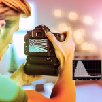 Eine Person hält eine Digitalkamera und betrachtet den Bildschirm, der ein Muster zur Belichtungswarnung anzeigt. Ein eingeblendetes Histogramm visualisiert die Helligkeitsverteilung des Bildes.
