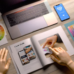 Ein Schreibtisch mit einem Laptop, Smartphone und verschiedenen Designmaterialien. Hände blättern durch ein offenes Buch, das App-Bildschirme und ein farbiges Designelement zeigt. Daneben liegen Farbräder und Farbfelder.