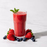 Ein Glas mit rotem Beerensmoothie, garniert mit Minze, umgeben von frischen Erdbeeren und Heidelbeeren auf einer weißen Marmoroberfläche.