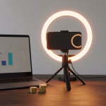 Ein Arbeitsplatz mit Laptop (Finanzgrafik), Goldmünzen und Smartphone mit Ringlicht, symbolisierend Online-Geldverdienen und Wachstum.