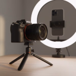Eine schwarze Kamera auf einem Stativ steht vor einem beleuchteten Ringlicht, in dem ein Smartphone befestigt ist.