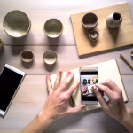 Hände fotografieren mit einem Smartphone ein Arrangement aus Keramikschalen und einer Pflanze auf einem Holztisch; auf dem Smartphone-Display ist ebenfalls ein Bild mit Händen beim Fotografieren zu sehen.