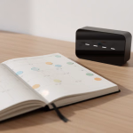 Ein aufgeklappter Planer mit bunten Kreisstickern auf den Daten liegt auf einem Holztisch neben einem schlanken schwarzen Gerät mit beleuchteten Strichen.