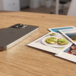 Ein dunkles Smartphone liegt auf einem Holztisch neben mehreren gestapelten Sofortbildern, die vielfältige Momente wie Avocado-Toast, eine sonnige Berglandschaft und eine gemütliche Decke zeigen.