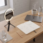 Ein Schreibtisch mit einem Smartphone auf einem Ringlicht-Stativ, einem offenen Notizbuch mit handgezeichneten Diagrammen, einem geschlossenen Laptop, einer dampfenden Kaffeetasse und Wasserflaschen.