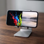 Ein Smartphone auf einem silbernen Ständer zeigt auf seinem Display zwei kontrastierende Bilder nebeneinander: links lebendige Farbspritzer auf schwarzem Grund und rechts eine ruhige, nebelverhangene Seenlandschaft.