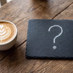 Eine Tasse Kaffee mit einem kunstvollen Blattmuster im Milchschaum steht neben einer schwarzen Schiefertafel mit einem großen, mit Kreide gezeichneten Fragezeichen auf einem rustikalen Holztisch.
