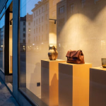 Die Dämmerung hüllt die Stadt in ein kühles Blau, während ein hell erleuchtetes Schaufenster einen warmen Kontrast bildet. Von einem dunklen Bürgersteig aus blickt man auf die Auslage eines Ladens, dessen Glasfront die Reflexionen der gegenüberliegenden Gebäude einfängt. Im Inneren präsentieren sich auf drei erhöhten, golden leuchtenden Podesten ausgewählte Objekte: eine kunstvolle, rustikale Vase, eine elegante braune Ledertasche und zwei schlichte Trinkgläser. Das sanfte Licht im Schaufenster hebt die Produkte hervor und lädt zum Verweilen ein, während das urbane Leben draußen in der Unschärfe verschwimmt. **Alternativtext:** Schaufenster eines Ladens in der Dämmerung, das einen Krug, eine braune Ledertasche und zwei Gläser auf hell beleuchteten Podesten zeigt.