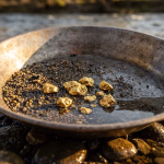 Eine Goldpfanne mit mehreren Goldnuggets und Kies am Ufer eines Flusses.