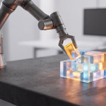 Ein Roboterarm platziert einen leuchtend orangefarbenen, durchsichtigen Baustein auf eine Struktur aus leuchtend blauen und orangefarbenen, durchsichtigen Blöcken, die auf einer dunklen Oberfläche stehen.