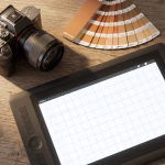 Eine digitale Kamera, ein Farbfächer und ein Grafiktablett mit Stift liegen auf einer Holzoberfläche.