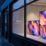 Moderne Fernsehgeräte in einem Schaufenster bei Nacht, die leuchtende, abstrakte Muster in Orange, Lila und Blau zeigen, mit einer nassen Straße und verschwommenen Stadtlichtern im Hintergrund.