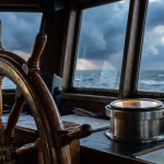 Ein hölzernes Steuerrad und ein Kompass in der Kabine eines Schiffes, mit stürmischer See und dunklen Wolken durch die Fenster sichtbar.