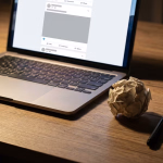 Laptop auf einem Holzschreibtisch, der einen Social-Media-Feed zeigt, daneben ein zerknüllter Papierball, ein Stift und eine Tasse, was auf die Arbeit an digitalen Inhalten hindeutet.