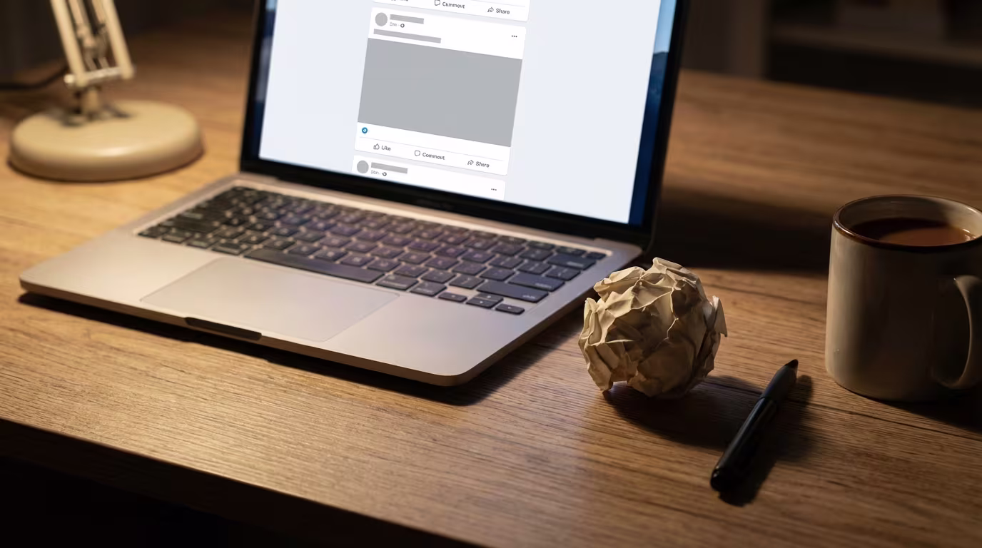 Laptop auf einem Holzschreibtisch, der einen Social-Media-Feed zeigt, daneben ein zerknüllter Papierball, ein Stift und eine Tasse, was auf die Arbeit an digitalen Inhalten hindeutet.